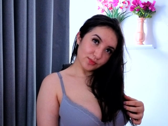 SusanMonroy with long black hair, light skin, and medium breasts, wearing a gray lace bra, slightly smiling, holding her hair, standing in front of gray curtains.