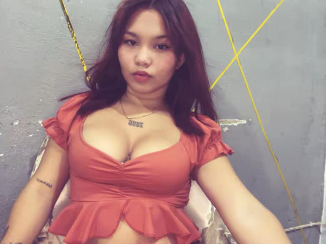 EstefyChaud with brown hair, wearing a red crop top that shows her big tits, has a tattoo on her right arm and a necklace with "ZEN" in cursive.