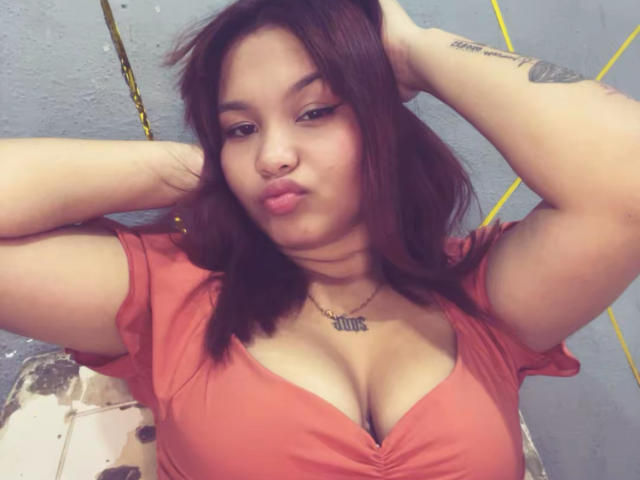 EstefyChaud with pouty lips, brown hair, wearing a red top, hands in hair, showing her big tits and tattooed arms, necklace with "Jewel" in the center.