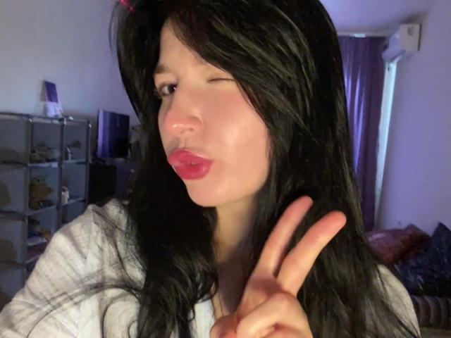 MertieDasinger with black hair, winking, and making a peace sign with her right hand, pouting pink lips, wearing a grey shirt, in a dimly lit room.