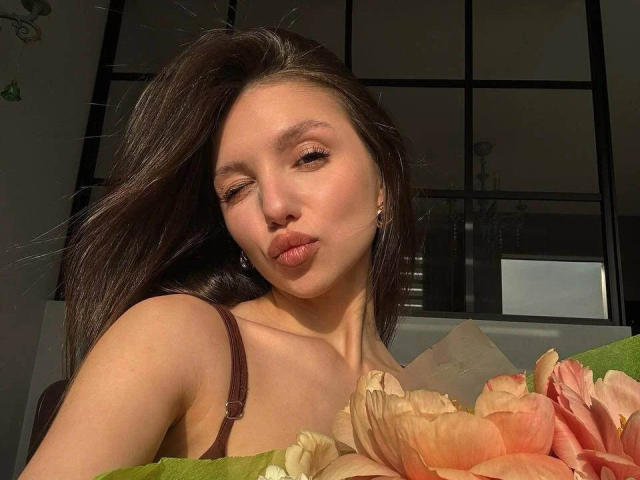 MaryroseCastine with pouty lips, brown eyes, long dark hair, brown strap dress, holding pink and orange flowers, sunlight on her face, earring visible.