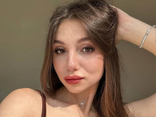 MaryroseCastine has brown hair, light skin, and wears a brown top with a silver bracelet. She's looking at the camera with a slight smile, her hand in her hair.