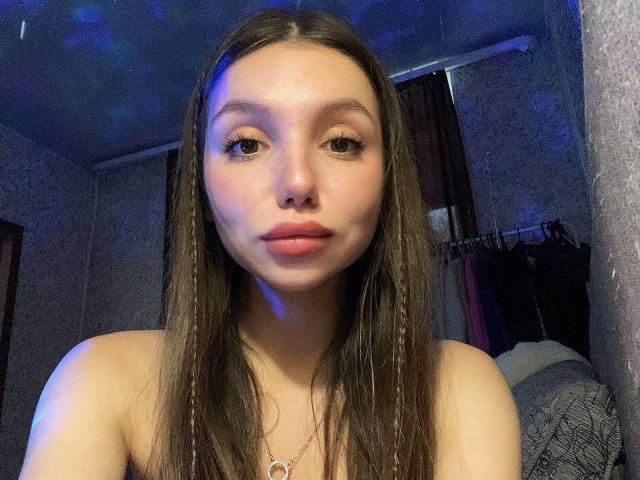MaryroseCastine with long brown hair, light skin, and pink lips, looking straight at the camera, topless, showing her small tits, wearing a heart necklace, in dim blue lighting.