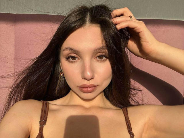 MaryroseCastine with long dark hair, brown eyes, and full lips, wearing a brown strap top, hand in hair, sunlight on face, and a ring on her finger.