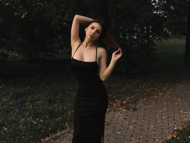 MaryroseCastine stands in a dark, wooded area, wearing a black, halter-neck dress, with one arm raised and the other touching her hair, showing her tits.