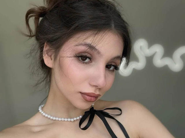 MaryroseCastine with dark hair in a messy bun, wearing a pearl necklace and black ribbon top, has pouty brown lips and long eyelashes, looking slightly to the side.