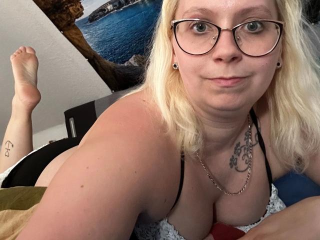 Stellaxx5, eine blonde Frau mit Brille, liegt im Bett, zeigt ihre Brüste in einem schwarzen BH, hat ein Tattoo auf der Brust und ein Tattoo am Knöchel, trägt eine Kette um den Hals, lächelt, die Beine sind angezogen, Foto..