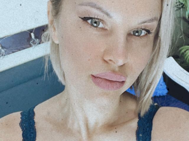 BelleLisaG with pouty pink lips, blonde hair, and black eyeliner, wearing a blue lace top, showing a clear, slightly freckled face and green eyes.