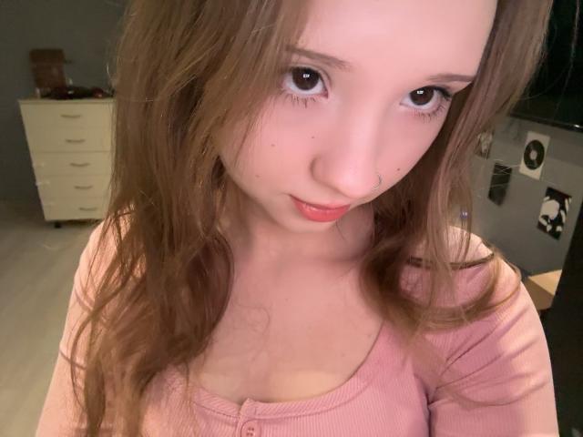 MarceneMalatesta, con cabello castaño largo, un pequeño aro en la nariz, una camisa rosa y una expresión adorable, mira hacia arriba con grandes ojos marrones, labios rosados ligeramente entreabiertos, mostrando sus pequeños pechos firmes..