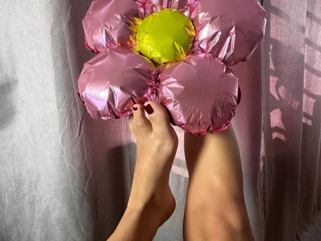 BalletSwan's feet, with pink nail polish, hold a shiny pink flower with a yellow center, against a gray curtain. Her toes are slightly curled, and sunlight highlights her skin.
