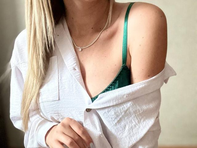 BalletSwan with blonde hair, green bra, and white shirt off-shoulder, showing bare shoulder and necklace. Boobs partially visible. Soft lighting, casual, sexy.
