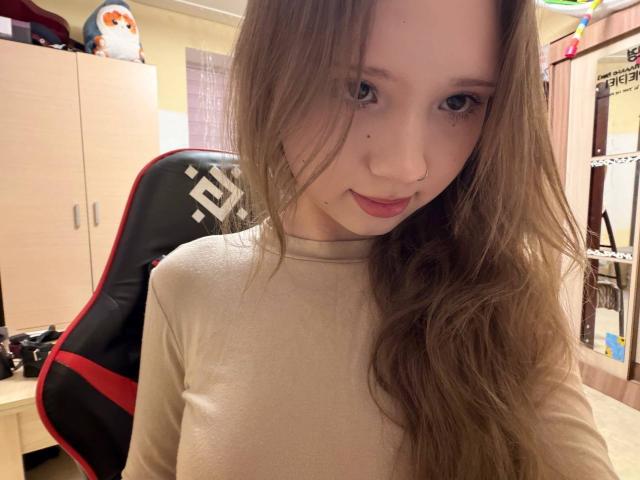 Marcene Malatesta, met lang bruin haar, gekleed in een beige shirt, zittend in een zwarte en rode gamingstoel, met een kleine moedervlek op haar linkerwang, met een subtiele glimlach..