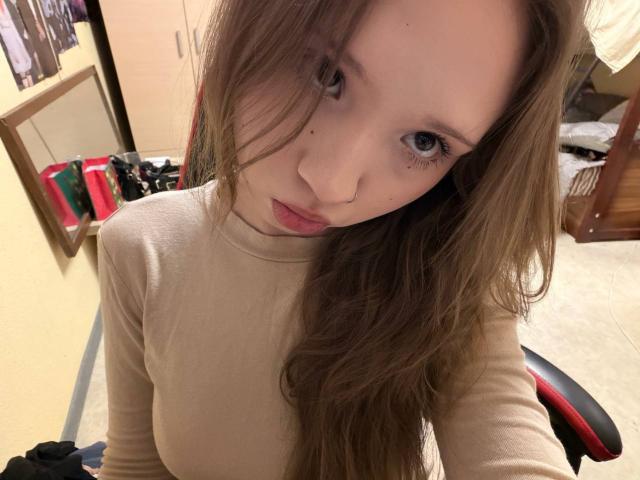 Marcene Malatesta, met lang bruin haar, blauwe ogen en roze lippen, draagt een beige shirt met lange mouwen en kijkt omhoog naar de camera, terwijl ze zit in een rode en zwarte stoel..