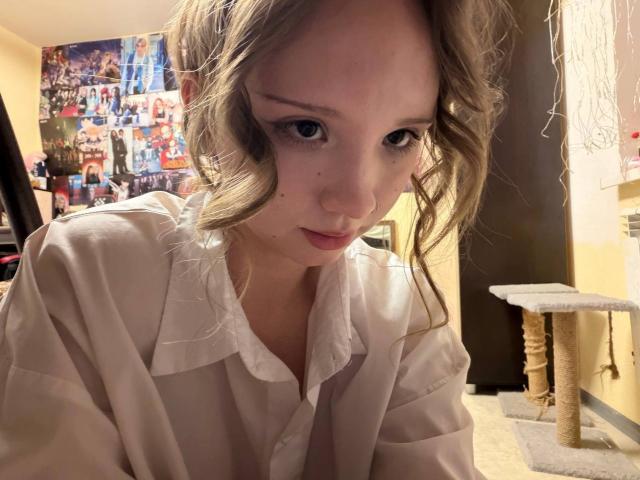 MarceneMalatesta, con cabello rubio ondulado, vestida con una camisa blanca de botones, mirando hacia abajo con una expresión ligeramente curiosa y somnolienta, y una pequeña peca en su mejilla izquierda..