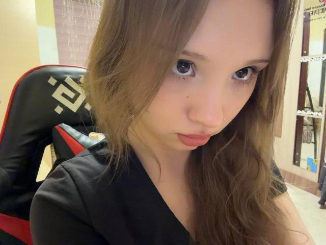 MarceneMalatesta with pouty lips, brown wavy hair, black shirt, and big blue eyes, sitting on a black and red gaming chair, showing a small beauty mark on her nose.