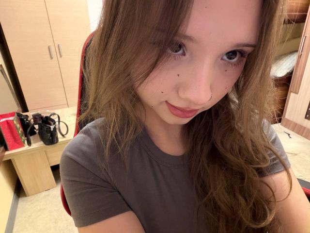 MarceneMalatesta, com cabelos castanhos longos, camisa cinza e sardas, sorrindo levemente, usando um piercing no nariz e olhando para a câmera. Seus seios não estão visíveis..