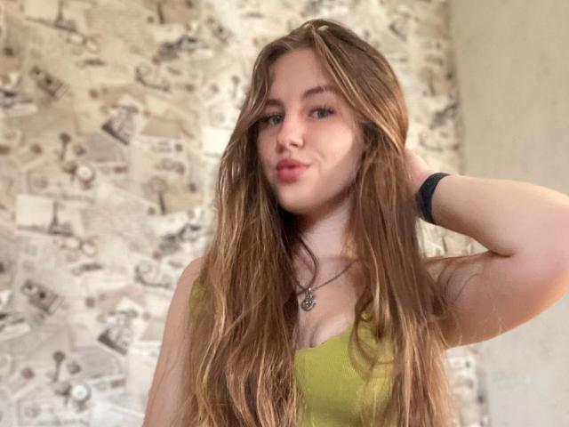 FroggyWonder con lunghi capelli castani, canotta verde e braccialetto nero, con un'espressione leggermente accigliata, una mano tra i capelli, che guarda la fotocamera, luce soffusa, casual, carina, con un leggero sorriso, rilassata..
