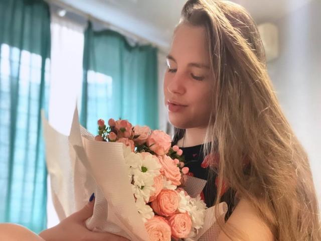 FroggyWonder com longos cabelos loiros, segurando um buquê de rosas rosa e brancas, olhando para baixo com um leve sorriso, em um quarto com iluminação suave e cortinas azuis..