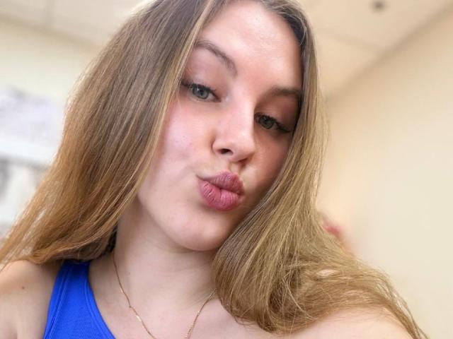 FroggyWonder mit hochgezogenen, rosafarbenen Lippen, einem blauen Oberteil, langen blonden Haaren, grünen Augen und einer goldenen Halskette, die ein Küsschen in einem Selfie gibt..