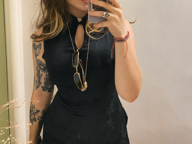 Mettis with wavy blonde hair, black sleeveless dress, tattooed arm, holding phone, gold necklace, glasses, red bracelet, taking mirror selfie, showing tits, in a mirror photo.