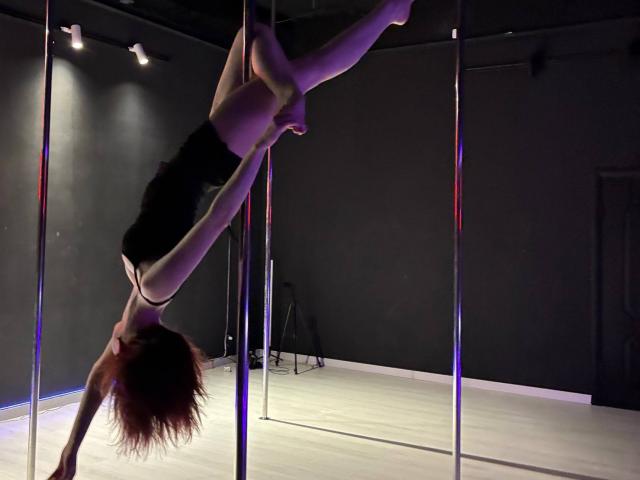 SarinaPadinha is upside down, hanging from a pole with her legs wrapped around it, wearing a black leotard, showing her ass and a small part of her tits.