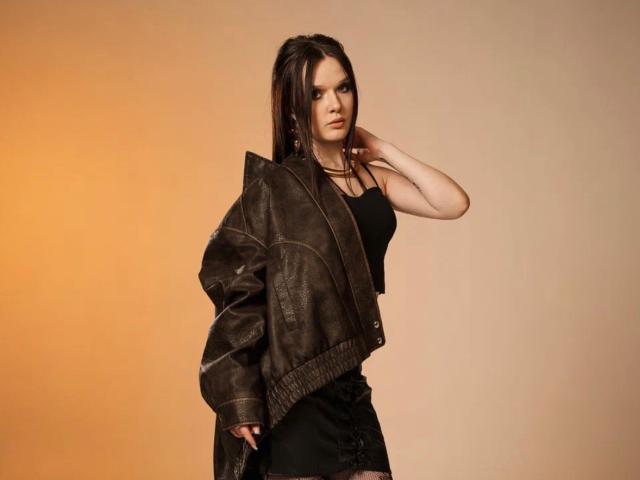 EsterGillom has dark brown hair, wearing a black tank top and black skirt, with a brown leather jacket draped over her shoulders, hand near her face.