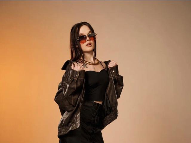 EsterGillom with dark brown hair, wearing red-tinted sunglasses, black strap top, and brown leather jacket, hands on jacket, small tattoo on chest, orange gradient background.