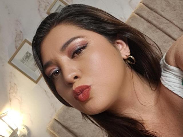 PaulaAnd with pouty red lips, brown eyes, shiny pink eyeshadow, and long brown hair, wearing gold hoop earrings, in a slightly angled selfie.