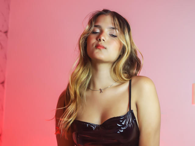 AbbySomer with wavy blonde hair, eyes closed, wearing a black vinyl top, slight pout, necklace with a small cross, red and pink light behind her.