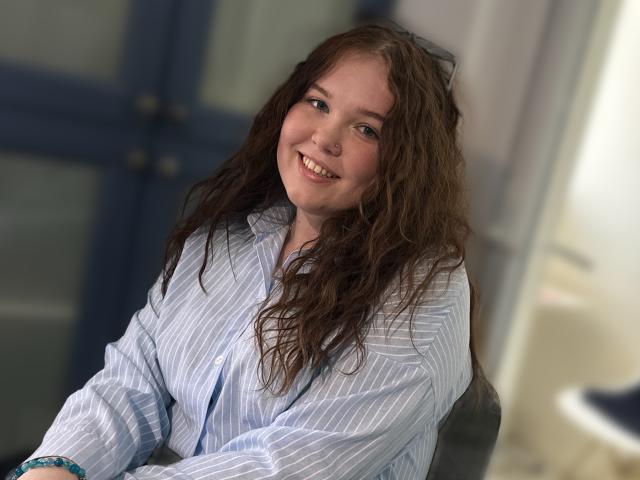 GorgeousVibe, met golvend bruin haar, lachend, gekleed in een blauw gestreept shirt, heeft een schattige neusring en een armband om haar rechterpols, en zit in een ontspannen houding..