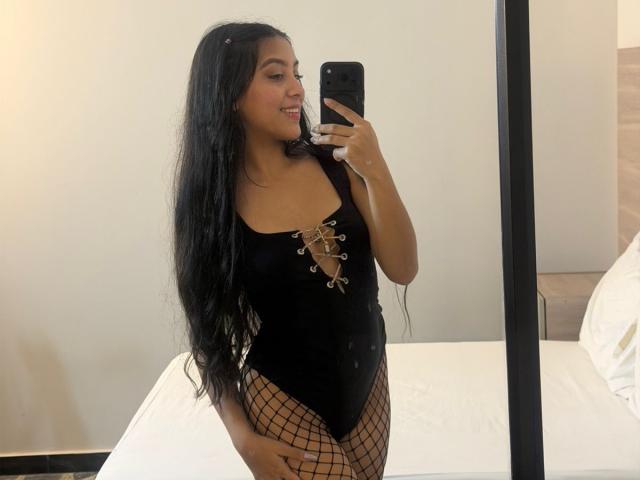EmmaEstreilla, avec ses longs cheveux noirs, portant une combinaison noire à lacets et des bas résille, se prend en photo devant un miroir, montrant ses fesses et ses seins, tout en souriant..