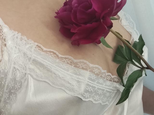 GladisBelles weißes Spitzenkleid mit tiefem Ausschnitt, das ihre Brust zeigt, und eine rote Rose, die sanft auf ihrer Brust platziert ist. Die grünen Blätter der Rose sorgen für einen Kontrast..