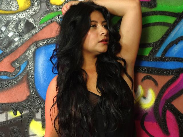 ArianaFoxxy with long, wavy black hair, arm raised, wearing a black top, standing against colorful graffiti, looking to the side with red lipstick. Her tits are visible.