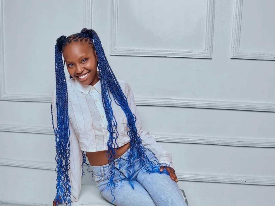 Una ragazza, SandNewbie, con lunghe trecce blu, sorridente, indossa una camicia bianca e jeans azzurri, seduta contro un muro bianco. Il seno non è visibile, il sedere non è visibile, i genitali non sono visibili..