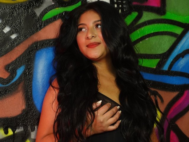 ArianaFoxxy with long black wavy hair, dark lipstick, and a black dress, covering her tits with her hand, smiling softly, standing against colorful graffiti.