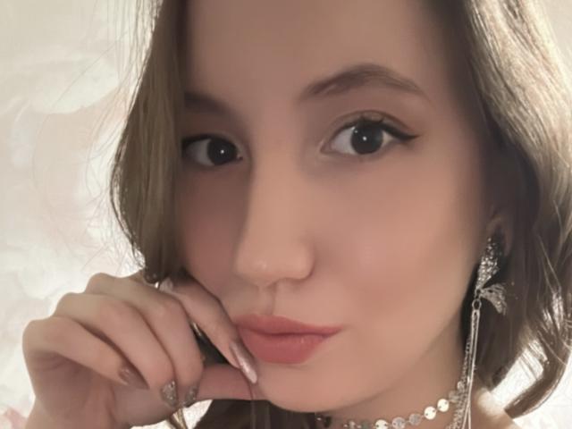 RoxyRoxie, avec ses lèvres roses et pulpeuses, ses cheveux bruns ondulés, son eye-liner discret, ses boucles d'oreilles en diamants et son collier en diamants, touchant son menton avec ses ongles manucurés, regardant l'appareil photo..