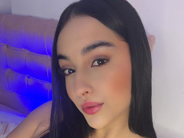 Emma Waltter, com cabelos longos e pretos, olhos castanhos e lábios rosados, usando delineador e com um leve sorriso, olhando para a câmera com uma expressão fofa e sensual..