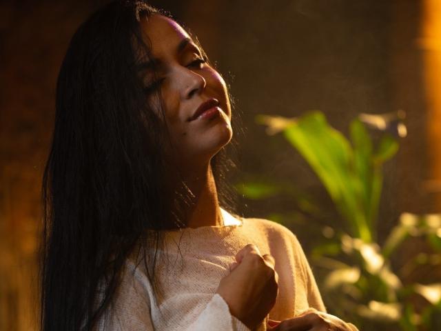 HikariChloe with dark wet hair, eyes closed, smiling softly, holding white shirt, warm light on face, green plant in blurred background, gentle and serene expression.