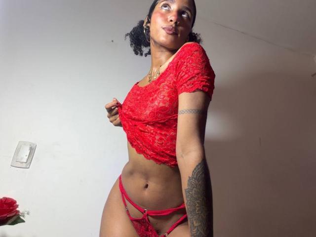 CinthiaWalker with dark curly hair, wearing a red lace top and matching red strappy panties, showing her tattooed arm and toned abs, looking up with a confident expression.