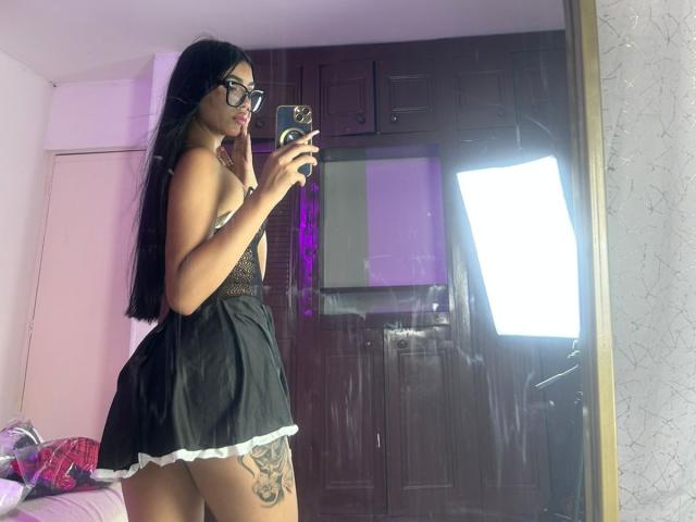 DulceNoir with long black hair, glasses, and a black lace lingerie set with white trim, showing a tattoo on her right thigh, taking a mirror selfie.
