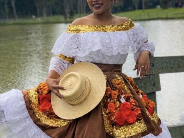 SophieSonyas in a white lace off-shoulder top, brown skirt with red and gold floral patterns, holding a straw hat, standing by a waterway, smiling, showing her tits.