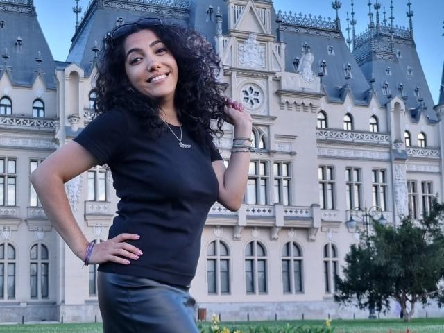 SexyMirra, con cabello negro rizado, sonriendo, vestida con una camiseta negra y pantalones de cuero negro, de pie frente a un edificio de estilo gótico, con las manos en las caderas, luciendo segura de sí misma..