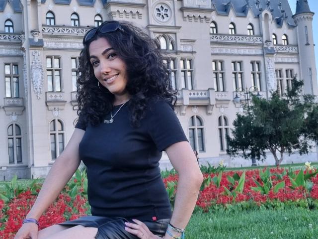 SexyMirra with curly black hair, black t-shirt, and black shorts, sitting on grass, smiling, with eyeglasses on head, hands on knees, red flowers and green plants behind.