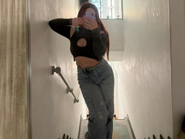 VicluX, avec ses longs cheveux roux, son haut noir découpé qui révèle ses seins, et son jean bleu déchiré, prend un selfie devant un miroir dans une pièce peu éclairée, tout en tenant un téléphone..