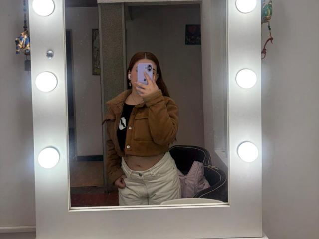 VicluX, avec ses cheveux bruns, portant une veste marron et un pantalon blanc taille haute, prend un selfie devant un miroir avec son téléphone, montrant son ventre plat et sa main posée sur sa hanche..