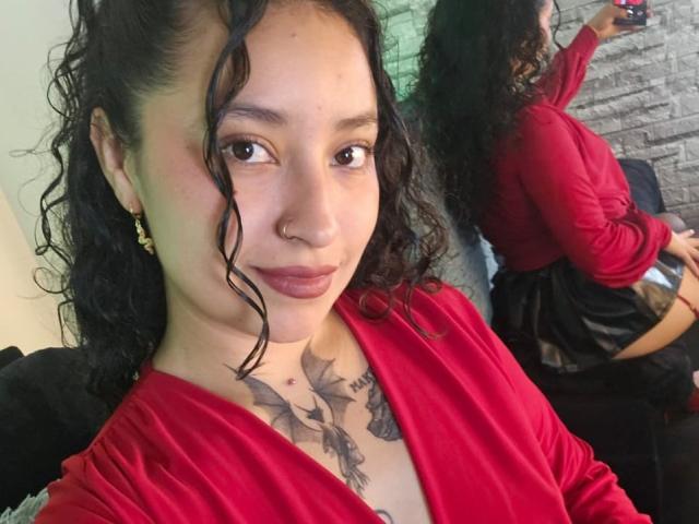 CarmilleZlata with curly black hair, nose ring, and red dress, showing tattooed chest, standing with back to camera, reflecting in mirror, holding phone. Boobs visible from side.