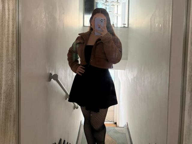 VicluX, con una giacca marrone, una gonna nera e calze a rete, è in piedi in un corridoio stretto, sta scattando un selfie allo specchio con il telefono, con le mani sui fianchi, mostrando una postura sicura..