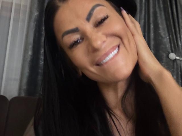 AndraD mit schwarzen Haaren, lächelnd, zwinkernd und mit der Hand auf ihre Wange gelegt. Sie hat dicke Augenbrauen, lange Wimpern und trägt ein schwarzes Oberteil. Ihre Zähne sind sichtbar..