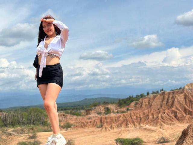 BibiSugarX with long black hair, white tied-up shirt, black mini-skirt, and white sneakers, standing on rocky terrain, hand shielding eyes, showing her ass and a tattoo on her side.