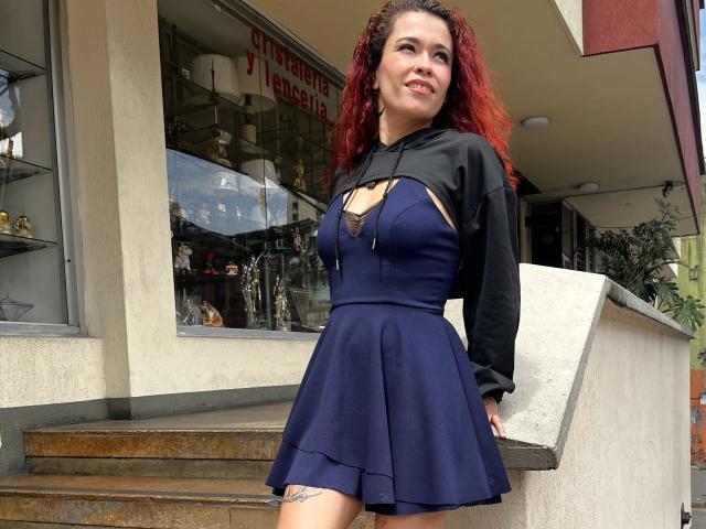 ArielleBrooks with red curly hair, wearing a black long-sleeve top with cut-out chest and a blue pleated skirt, standing on stairs, smiling, showing a tattoo on her thigh.